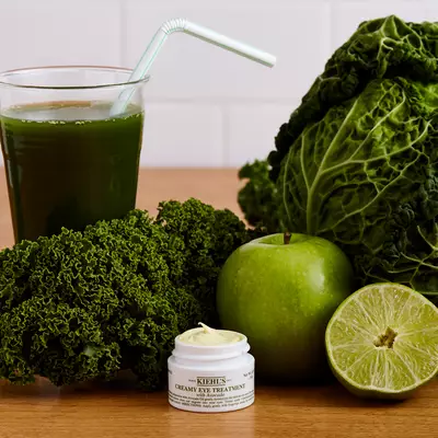 Gruner Smoothie mit Strohhalm, frischem Grunkohl, Apfel, Limette und einer Kiehls Augencreme auf einem Holztisch.
