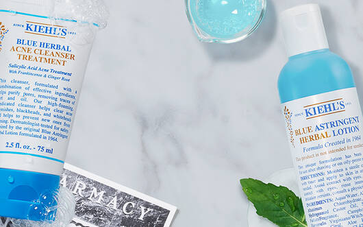 Alt-Text: Eine flache Anordnung von Kiehl's Hautpflegeprodukten auf einem marmorierten Hintergrund, einschließlich einer Tube mit Gesichtscreme, einer Flasche mit Lotion und frischen Krautern.