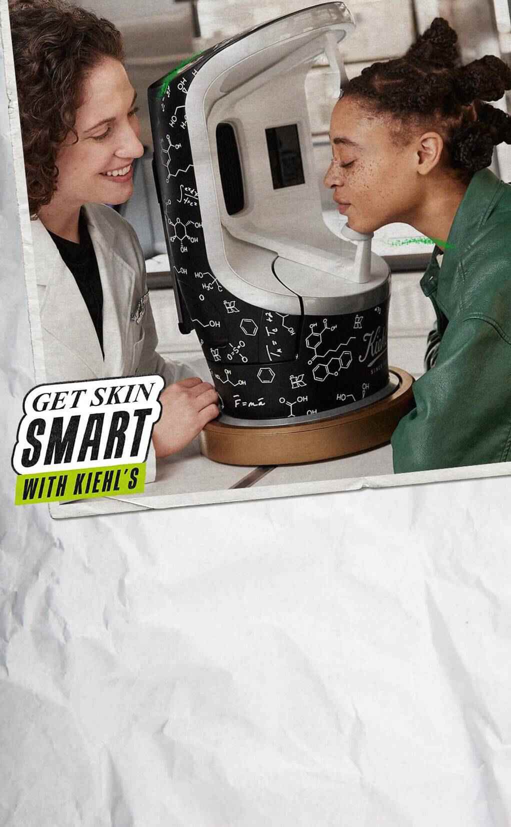Zwei Personen stehen vor einem Kiehl's Hautanalysegerat. Eine Person lachelt, wahrend die andere das Gerat anblickt. Das Gerat ist mit chemischen Formeln und dem Kiehl's Logo verziert.