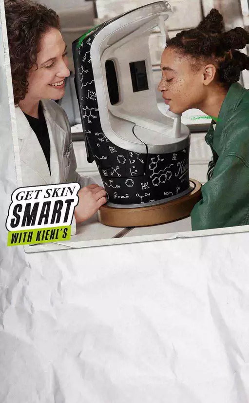 Zwei Personen stehen vor einem Kiehl's Hautanalysegerat. Eine Person lachelt, wahrend die andere das Gerat anblickt. Das Gerat ist mit chemischen Formeln und dem Kiehl's Logo verziert.