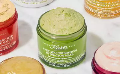 Grune Avocado-Nahrungshydrationsmaske von Kiehl's, umgeben von verschiedenen anderen Hautpflegeprodukten auf einem Marmoruntergrund.