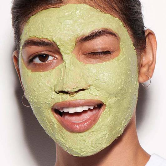 Eine Frau mit gruner Gesichtsmaske, einem Zwinkern und einem breiten Lacheln blickt in die Kamera.