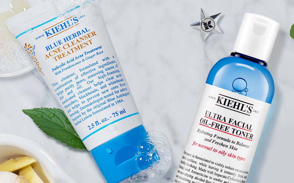 Produktanordnung auf marmoriertem Hintergrund, einschließlich Kiehls-Flaschchen mit Gesichtstoner und Feuchtigkeitscreme, umgeben von Krautern und weiteren Schonheitsprodukten.