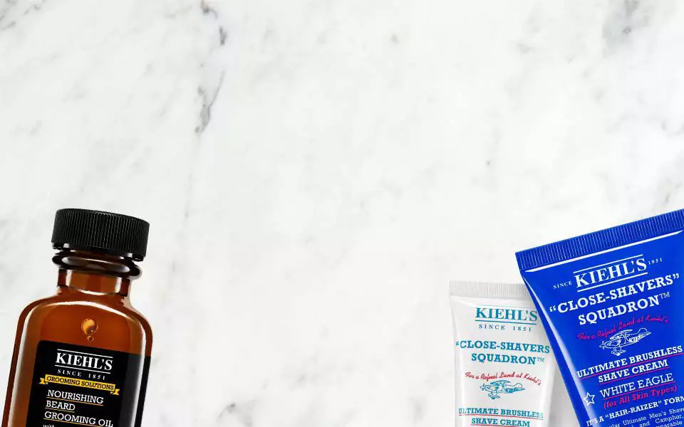 Alt-Text: Eine Anordnung von Kiehl’s Produkten auf einem marmorierten Hintergrund, einschließlich eines Gesichtsols, einer Rasiercreme und einer Aftershave-Creme.