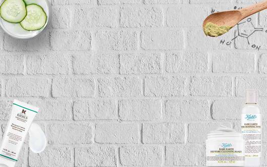 Ein grauer Backstein-Hintergrund mit Hautpflegeprodukten, einer Schussel mit Gurken und einem Holzloffel, der eine grune Substanz halt.
