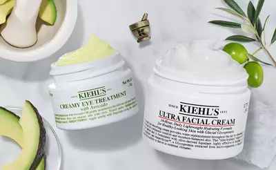 Zwei Kiehl's Hautpflegecremes: Avocado-Augenbehandlung und Ultra Gesichtscreme, umgeben von Avocado und Oliven.