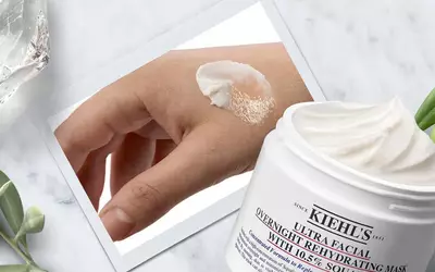 Ein Hand mit feuchtigkeitsspendender Gesichtsmaske von Kiehl's, daneben die offene Verpackung auf einer grunen Unterlage.