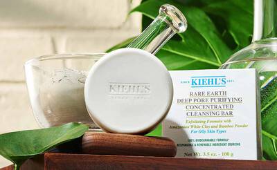 Ein Kiehl's Reinigungssubstanz in einem weißen Behalter, umgeben von grunen Pflanzen und einem Papieretikett.