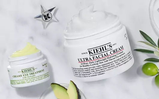 Alt-Text: Eine flache Anordnung von Kiehl's Hautpflegeprodukten auf einem marmorierten Hintergrund, umgeben von frischen Pflanzen und Zutaten.