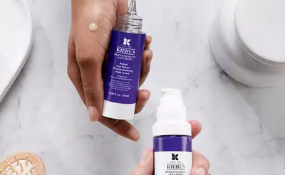 Hande halten Kiehl's Hautpflegeprodukte: ein Serum und eine Creme auf marmoriertem Tisch.