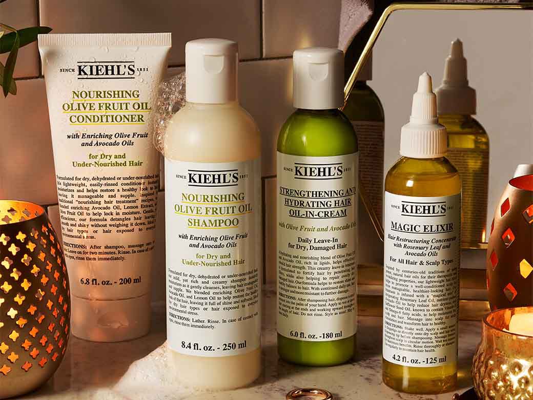 Eine Reihe von Kiehl's Haarpflegeprodukten (Shampoo, Spulung, Ole) mit Oliven- und Avocadool auf einem Waschtisch arrangiert.