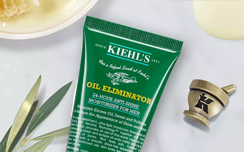 Bildbeschreibung: Eine grune Tube mit dem Text "Oil Eliminator" von Kiehl's liegt auf einer marmorartigen Oberflache, umgeben von kosmetischen Produkten und einer kleinen Schale mit einer unbekannten Substanz.