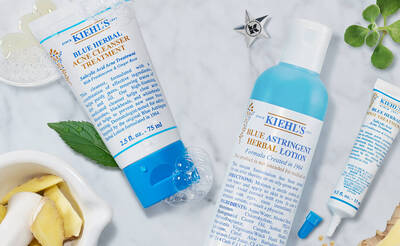 Kiehls Produkte: Blaues Krauter-Akne-Reinigungsgel und astringierende Lotion auf einem Tisch mit Krautern und Zutaten.