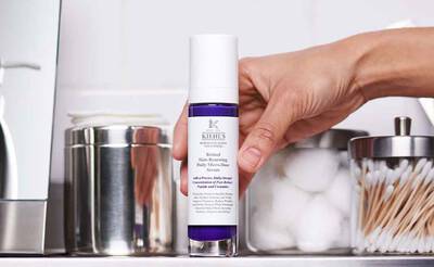 Un flacon de serum de la marque Kiehl's, tenu par une main, avec des accessoires de salle de bain en arriere-plan.