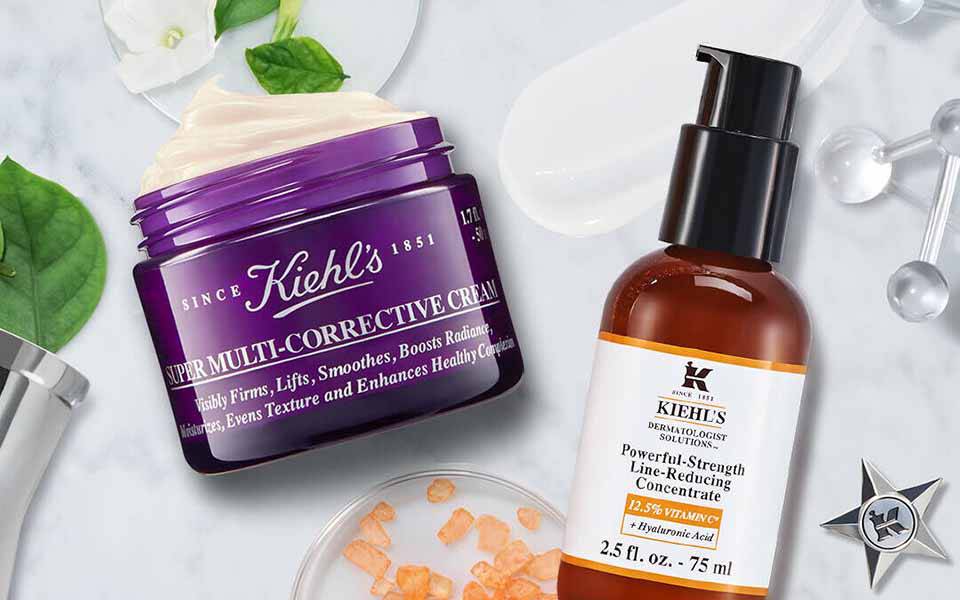 Alt-Text: Eine Szene mit verschiedenen Hautpflegeprodukten, darunter eine lila Creme von Kiehl's und ein braunes Serum von Kiehl's, umgeben von grunen Blattern und weiteren Beauty-Requisiten auf einer marmorierten Oberflache.