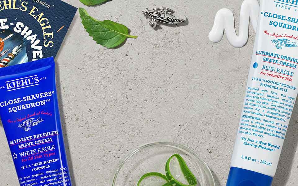 Alt-Text: Verschiedene Kiehl's Produkte fur die Rasur, darunter Rasiercreme und Aftershave, auf einem hellen Hintergrund mit frischen Krautern und einem Rasierer.