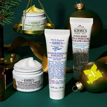Eine festliche Anordnung von Kiehl’s Produkten, darunter Creamy Eye Treatment auf goldenen Waagschalen, Ultra Facial Cream, Ultimate Strength Hand Salve und Ultra Body Mega Moisture Squalane Cream, umgeben von grunen Ornamenten, Tannenzweigen und goldenen Bandern auf einem dunkelgrunen Hintergrund.