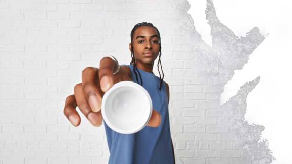 Eine Person in einem blauen armellosen Shirt halt eine kleine weiße Schale in Richtung Kamera, vor einer strukturierten weißen Wand.