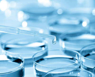 A close-up view of a pipette with a drop of liquid poised above several transparent petri dishes, all set against a blue background.
