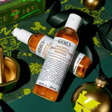 Kiehl’s Calendula Herbal-Extract Toner und Serum, arrangiert mit goldenen Weihnachtsornamenten und grunem Band auf einem festlichen, dunkelgrunen Hintergrund.